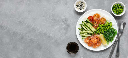 Salad of rice, salmon, cucumber, chuka seaweed, he beans, cherry tomatoes and sesame seeds in a white plate on a gray concrete table. Healthy food. Top view, bannerの写真素材