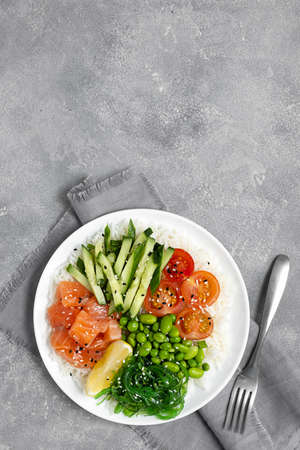 Salad of rice, salmon, cucumber, chuka seaweed, he beans, cherry tomatoes and sesame seeds in a white plate on a gray concrete table. Healthy food. Top viewの写真素材