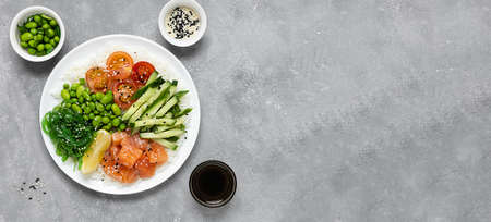 Poke salad of rice, salmon, cucumber, chuka seaweed, he beans, cherry tomatoes and sesame seeds in a white plate on a gray concrete table. Healthy food. Top view. Bannerの写真素材
