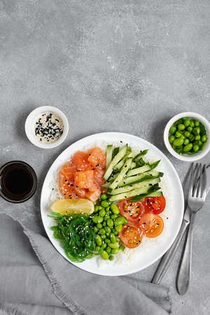 Salad of rice, salmon, cucumber, chuka seaweed, he beans, cherry tomatoes and sesame seeds in a white plate on a gray concrete table. Healthy food. Top viewの写真素材
