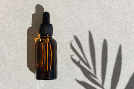 Bottle of dark amber glass for essential oil, cream, lotion or serum on a gray background with a shadow of tropical palm leaves. Presentation of a cosmetic product. Layout of the beauty salon brandingの写真素材