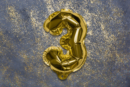 The number of the balloon made of golden foil, the number three on a gray background with sequins. Birthday greeting card with inscription 3. Top view. Numerical digit, Celebration event, template.の写真素材