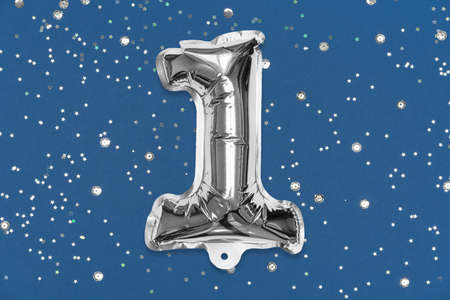 Silver foil balloon number, digit one on a blue background with sequins. Birthday greeting card with inscription 1. Top view. Numerical digit. Celebration event, template.の写真素材