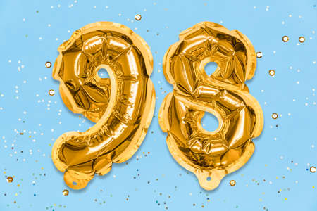 The number of the balloon made of golden foil, the number ninety-eight on a blue background with sequins. Birthday greeting card with inscription 98. Numerical digit. Celebration event template.の写真素材