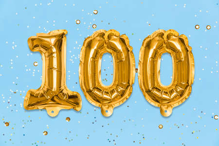 The number of the balloon made of golden foil, the number one hundred on a blue background with sequins. Birthday greeting card with inscription 100. Anniversary concept. Celebration event template.の写真素材