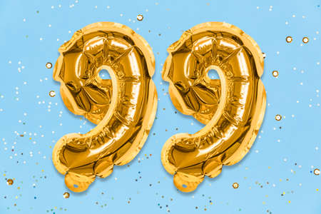 The number of the balloon made of golden foil, the number ninety-nine on a blue background with sequins. Birthday greeting card with inscription 99. Numerical digit. Celebration event template.の写真素材