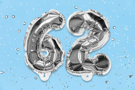Silver foil balloon number, digit sixty two on a blue background with sequins. Birthday greeting card with inscription 62. Anniversary concept. Top view. Numerical digit. Celebration event, template.の写真素材