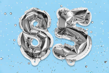 Silver foil balloon number, digit eighty five on a blue background with sequins. Birthday greeting card with inscription 85. Anniversary concept. Numerical digit. Celebration event template.の写真素材