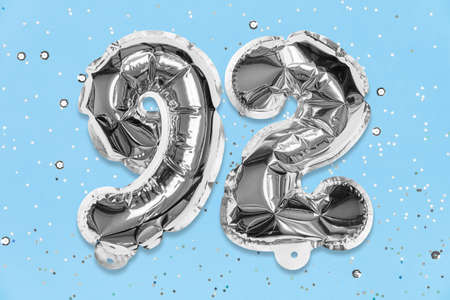 Silver foil balloon number, digit ninety two on a blue background with sequins. Birthday greeting card with inscription 92. Top view. Numerical digit. Celebration event, template.の写真素材