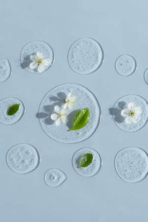 Drops of transparent cosmetic gel or serum with white flowers and green leaves on a blue background. The concept of natural cosmetics.の写真素材