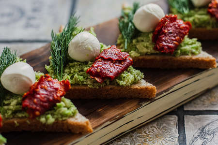 Mini triangle canapes bruschetta with avocado, mozarella and dried tomatos on the rustic board on the vintage tiled background.の写真素材