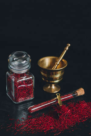 Raw organic red dried saffron spice on wooden background in vintage metal brass mortar with pestle, glass jar and tubeの写真素材