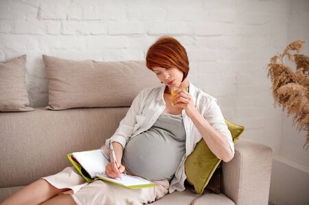 Pregnant woman drinks juice and writes plans in a diary while lying on a sofa. Selective focusの写真素材