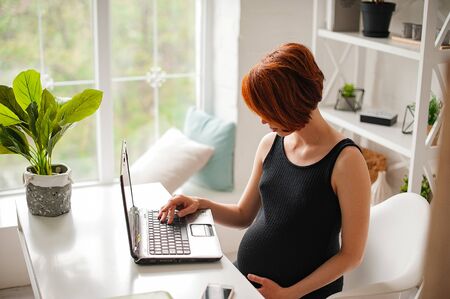 Pregnant woman working on laptop. Image of pregnant business woman typing something on laptop while sitting at her working place in officeの写真素材