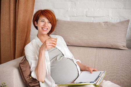 Pregnant woman drinks juice and writes plans in a diary while lying on a sofa. Selective focusの写真素材