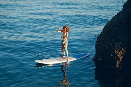 Young women Having Fun Stand Up Paddling in the sea. SUP. Red hair girl Training on Paddle Board near the rocks.の写真素材