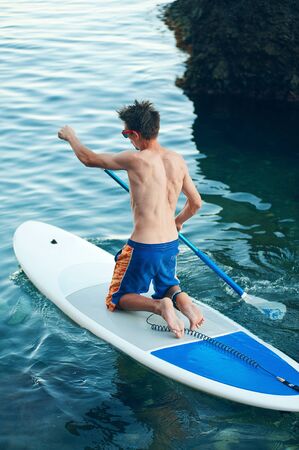 Young Man Having Fun Stand Up Paddling in the sea. SUP. Guy Training in the morning on Paddle Board near the rocks.の写真素材