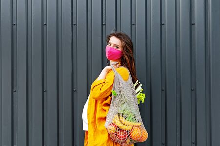 Pregnant woman in a protective mask and yellow clothes with an eco bag with fruits and vegetables on...