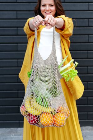 Pregnant woman in yellow clothes with an eco bag with fruits and vegetables on a black wall background. Plastic free concept Sustainable lifestyle.の写真素材