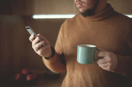 Young man drinks coffee and looks into a smartphone in the kitchenの写真素材