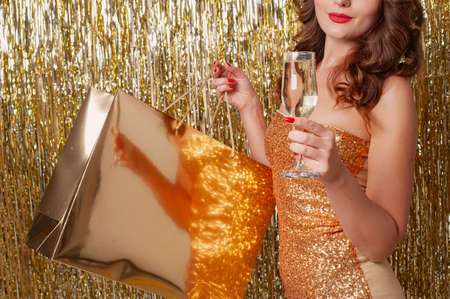 Woman in fashion clothing with a golden paper bag drinks champagne on a shiny gold backgroundの写真素材