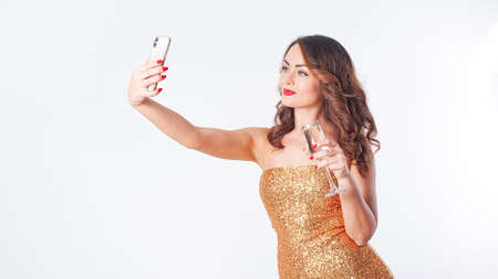 Young woman drinks champagne, makes a selfie on a smartphone, having fun on a white backgroundの写真素材