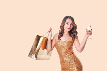 Woman in fashion clothing with a gold paper bag and a glass of sparkling wine on a beige backgroundの写真素材