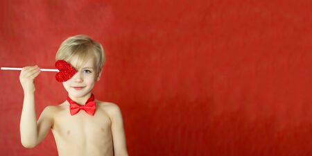 pretty cupid boy with a triple smile, bow tie and heart arrow isolated on red background. Copyspace for text. Valentines day concept. Bannerの写真素材