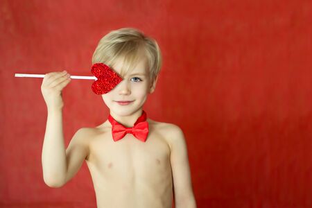 pretty cupid boy with a triple smile, bow tie and heart arrow isolated on red background. Copyspace...