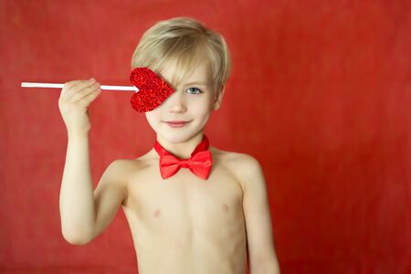 pretty cupid boy with a triple smile, bow tie and heart arrow isolated on red background. Copyspace for text. Valentines day concept.の写真素材