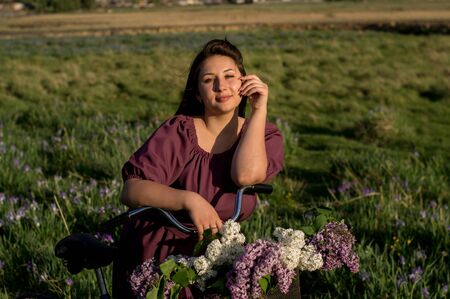 A beautiful young Caucasian smiling happy girl in purple dress in a wreath and with a bicycle a basket of blooming lilac in the countryside. Model plus size. Spring, rustic style. Circlet of flowers.の写真素材