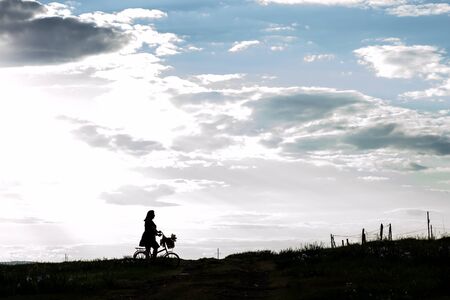 Silhouette beautiful young girl on bicycle a basket of blooming lilac in the countryside against a dramatic sky . Model plus size. Spring, rustic style.の写真素材