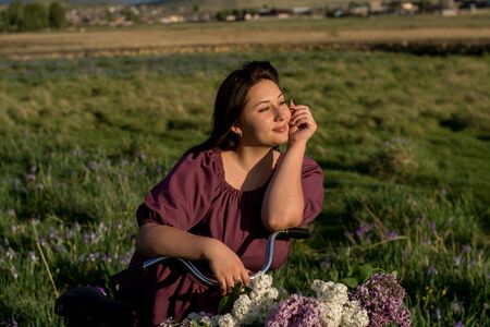 A beautiful young Caucasian smiling happy girl in purple dress in a wreath and with a bicycle a basket of blooming lilac in the countryside. Model plus size. Spring, rustic style. Circlet of flowers.の写真素材