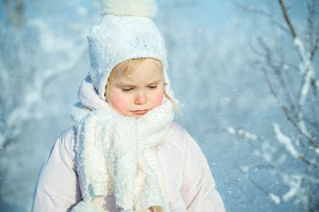 A child in a warm white hat and scarf plays with snow while walking in the winter forest, snowfall, winter magic, bright emotion, protecting the skin from frostの写真素材