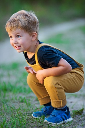 Cute little boy in casual clothes sitting on his haunches and smiling on summer day in countrysideの写真素材