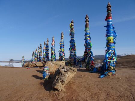 a row of ritual Buryat pillars wrapped with multicolored ribbons on a background of sand snow and blue skyの写真素材