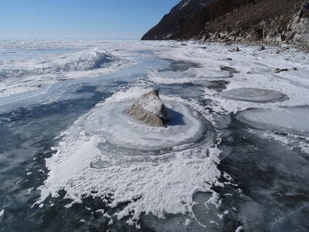 strangely frozen ice forms bright circles around large boulders near the rocky shore against the background of sparkling ice hummocks and blue skyの写真素材
