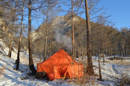 A red tent stands in the forest on the snow against the blue sky and mountains in winter smoke comes from the chimneyの写真素材