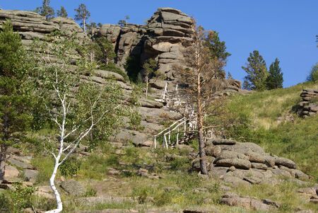 a staircase made of wood rises to a high rock of unusual shape, trees, birch and pines grow around itの写真素材