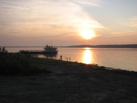 The sun sets over a large river. There is an old ferry near the shoreの写真素材