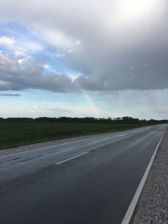 Above the wet, gray paved road with white markings, the sky is overcast and overcast. Rainbows and sunlight can be seen among the cloudsの写真素材