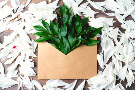 white petals on wooden background. open envelope of craft paper with buds and leaves of peonies in it. flat lay, top viewの写真素材