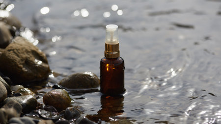 An amber cosmetic spray bottle placed in shallow river water with smooth stones, perfect for natural and eco-friendly product branding.の写真素材