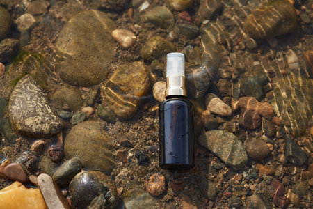 Dark cosmetic spray bottle with a golden cap placed on riverbed stones in clear shallow water. Natural skincare and wellness product conceptの写真素材