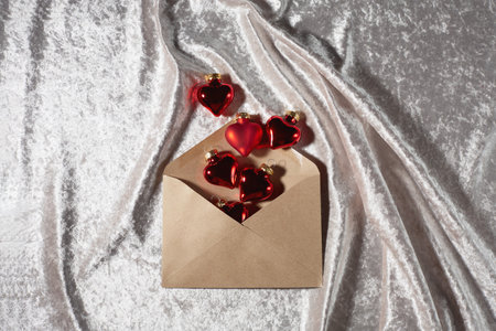 Red heart decorations are spilling from a brown envelope placed on shimmering silver fabric, creating a romantic and festive atmosphere.の写真素材