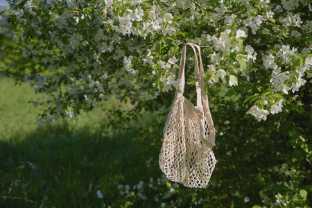 A lightweight tote bag hangs from a branch adorned with white flowers, surrounded by lush greenery...