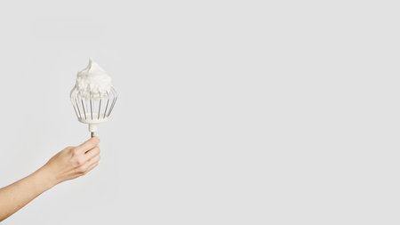 Panoramic banner with space for text. Hand holds a whisk covered in fluffy whipped cream, showing...