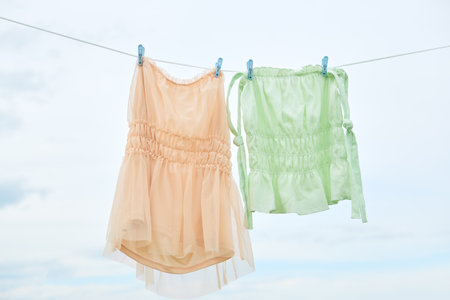 Peach and green sleeveless blouses hanging on a clothesline with blue clips against cloudy sky. Studio fashion photographyの写真素材