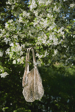 A woven bag is suspended from a branch adorned with vibrant white blooms, capturing the essence of spring in a serene outdoor setting.の写真素材