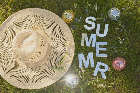 Wide overhead summer composition featuring a woven straw hat on green grass beside reflective decorative spheres, paper letters spelling summer, small wildflowers, and bright natural